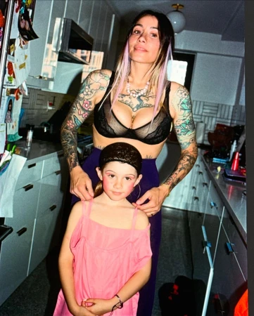 Woman and child pose indoors. Woman has tattoos and a black bra. The child wears pink dress. The scene is domestic and personal.