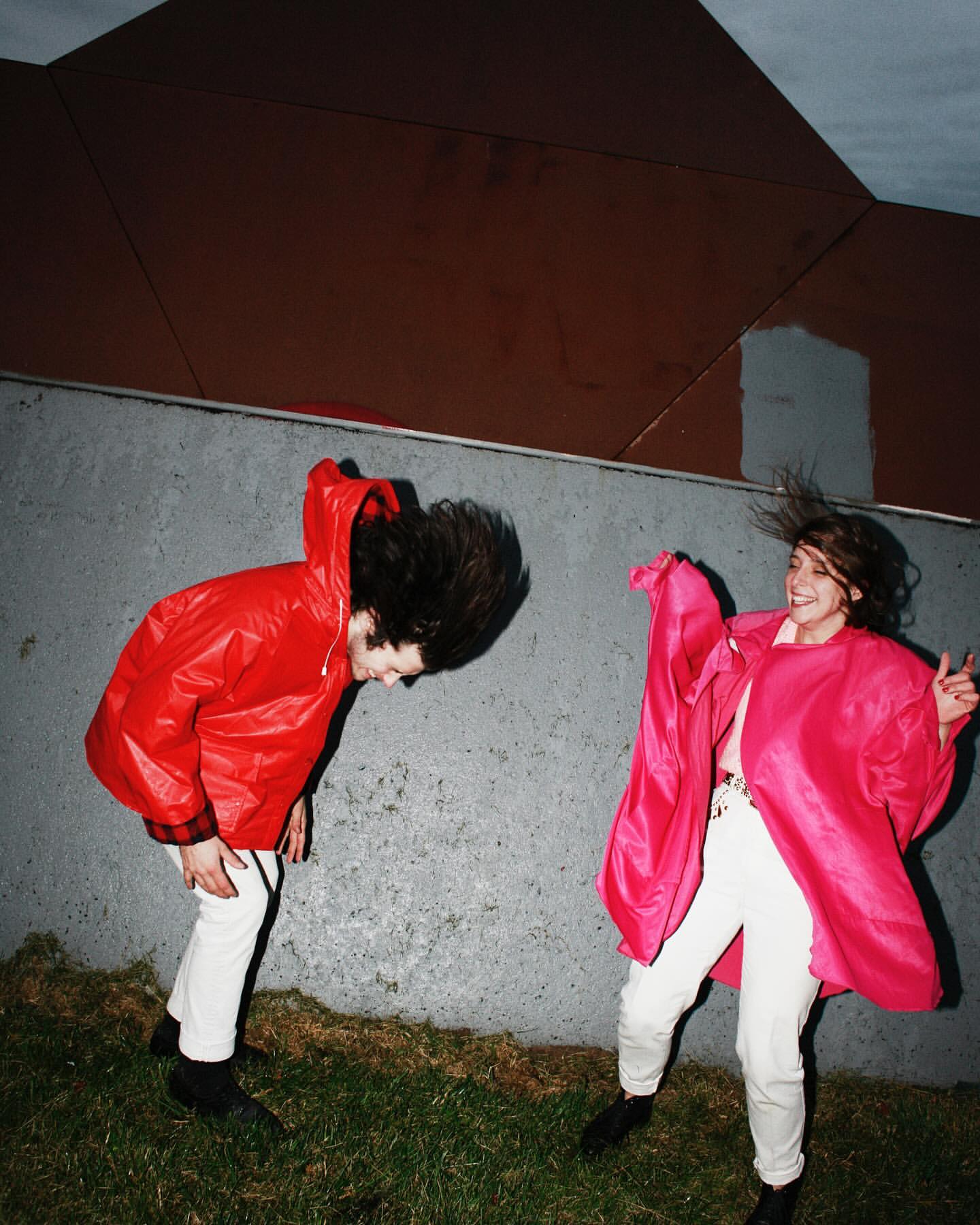 Two people in bright pink and red raincoats pose outdoors with windswept hair against a concrete and geometric background on a green lawn.