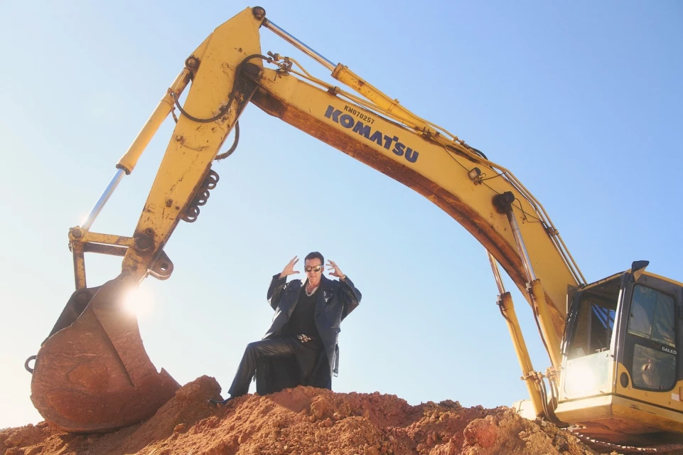 Adult male in black clothing poses on a mound of dirt in front of yellow Komatsu excavator under a clear blue sky in a medium shot.