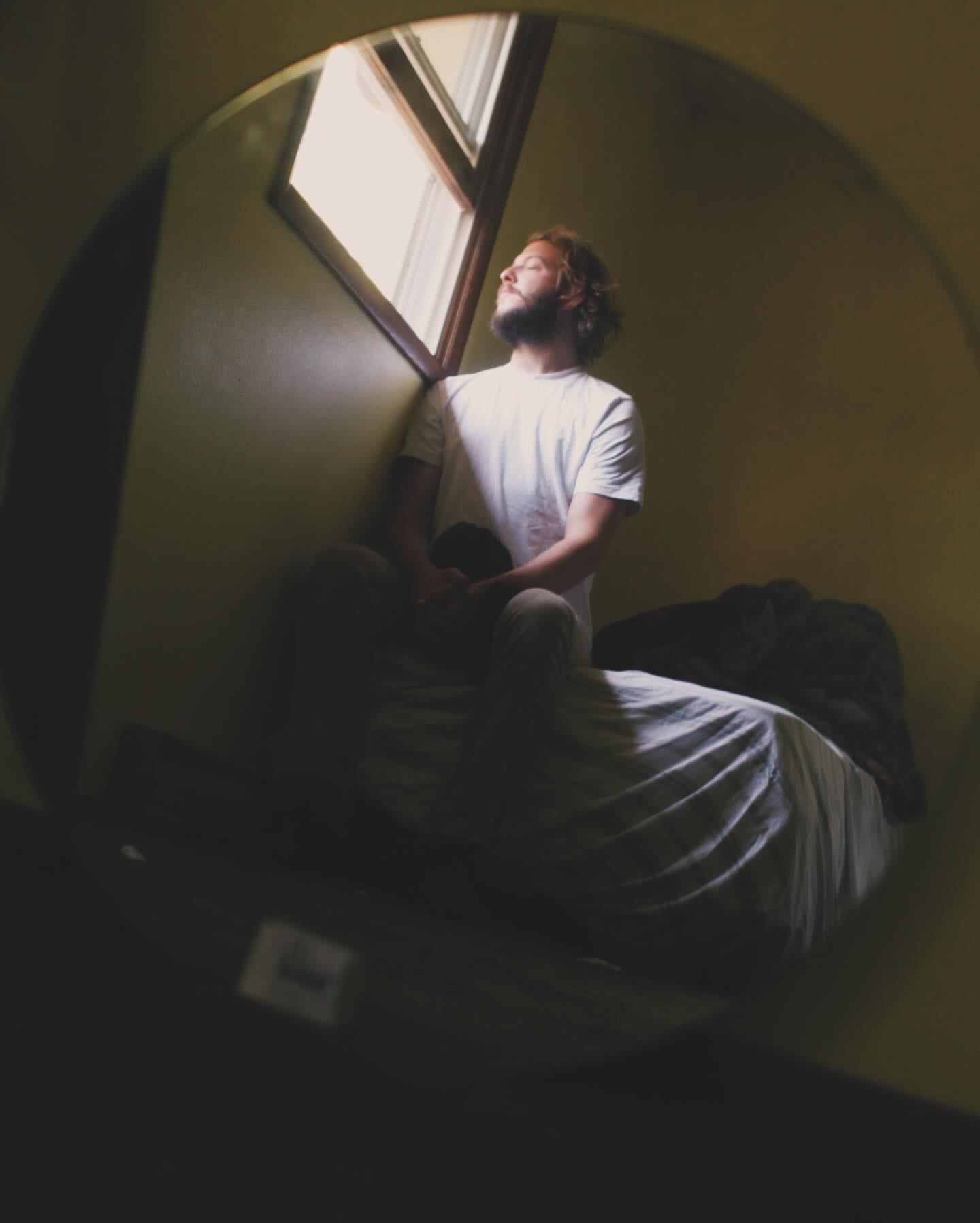 Indoor shot of an adult male in a white t-shirt seated on a bed in front of a window with natural light, captured in a round frame.