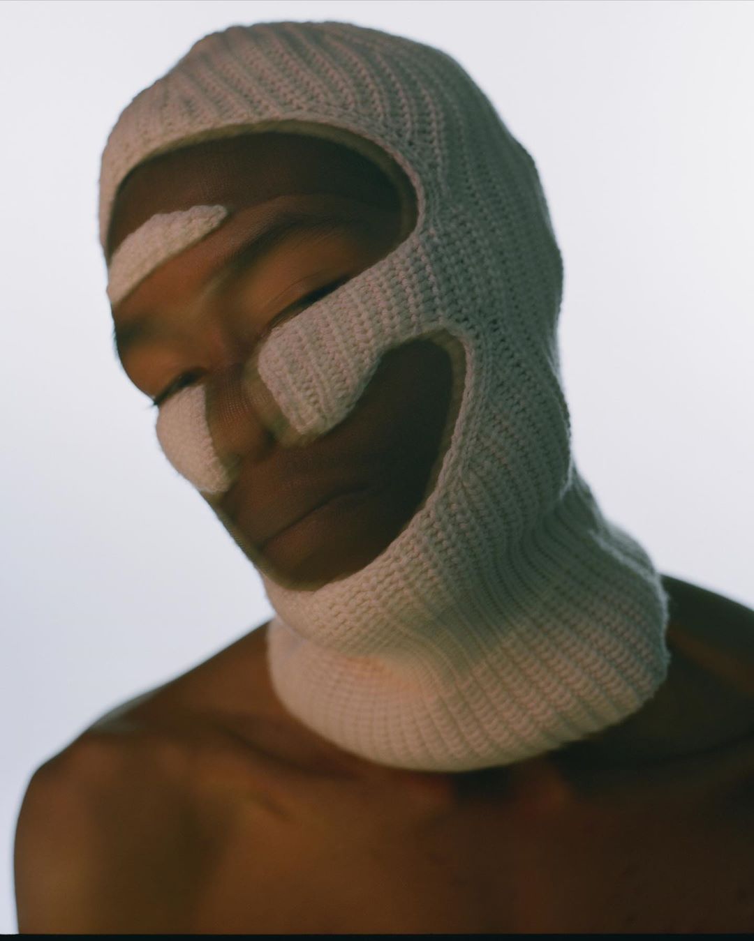 An adult with neutral expression wearing a textured white balaclava and white paint on the face in a close-up studio portrait.
