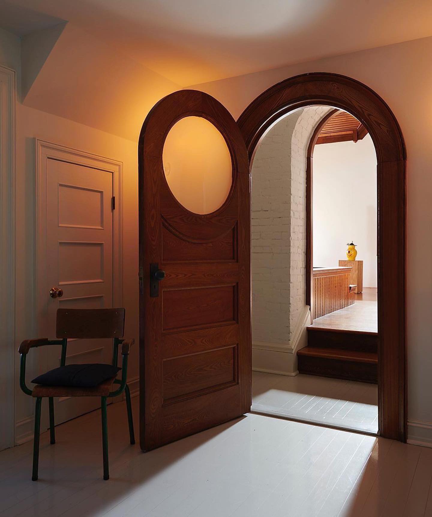 Arched wooden doorway frames view of a staircase; a door with a circular window is slightly ajar, with chair in foreground.