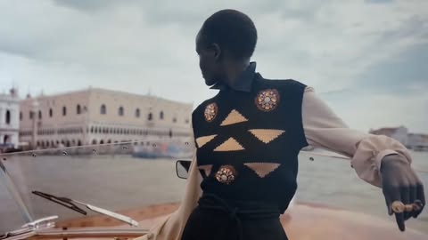 A dark-skinned adult female wearing a black vest in a car, looking at a building across the water with a cloudy sky in the background.