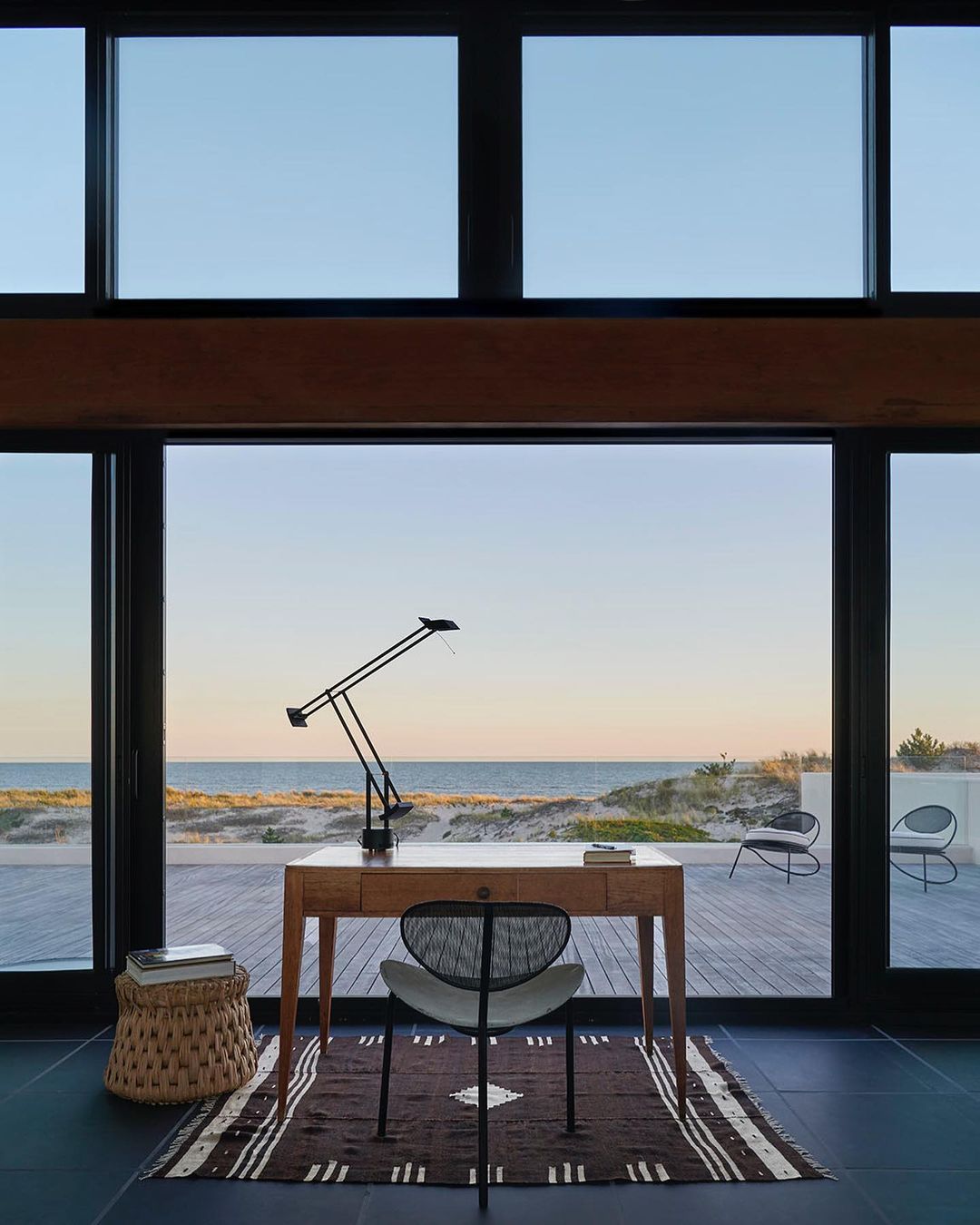 Home office interior with wooden desk, black chair, table lamp, and view of the ocean through the large windows, with a brown rug on the floor.