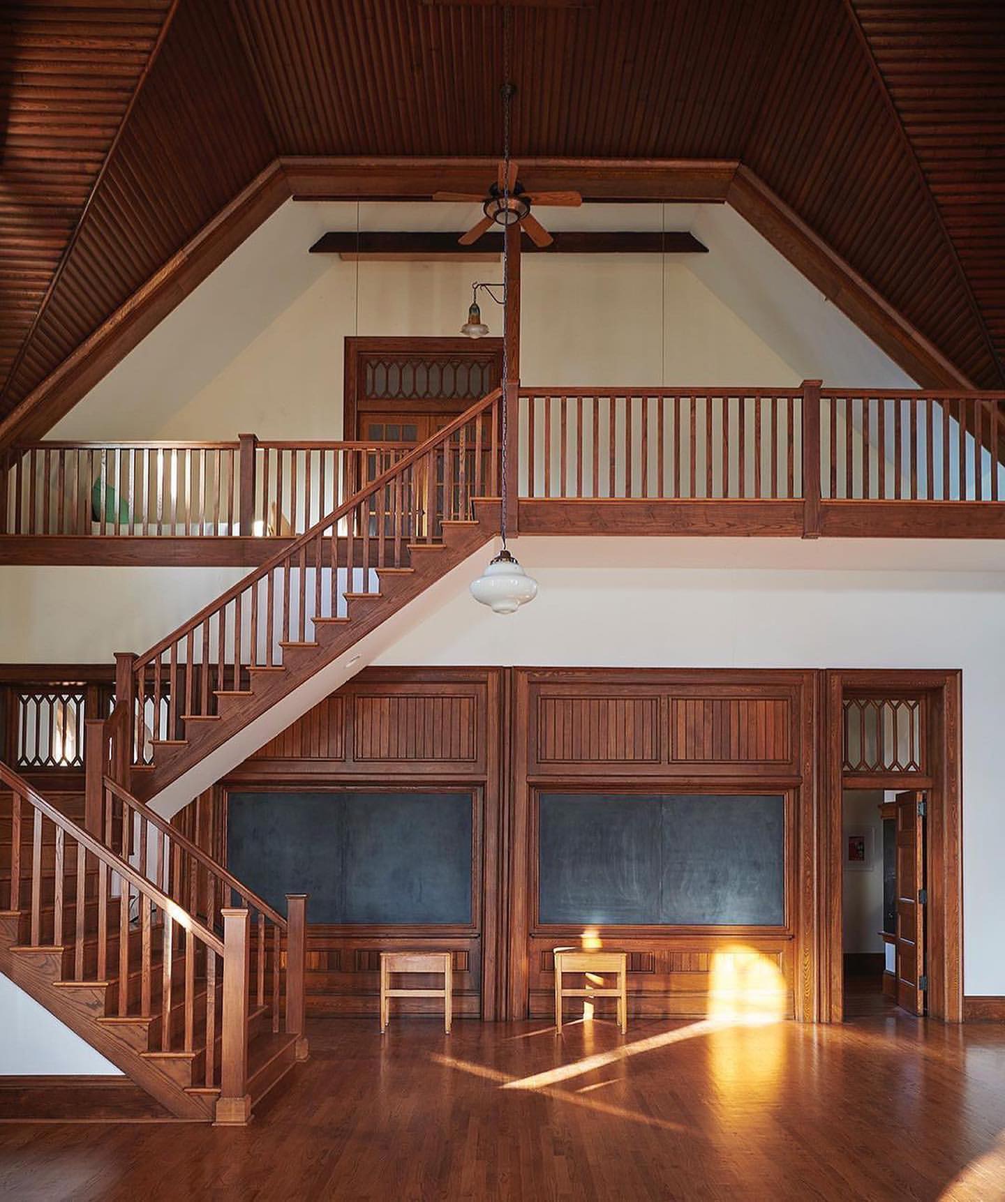 Interior view of a two-story space with a staircase, wood-paneled walls, and natural light streaming in through the doorway onto the wooden floor.