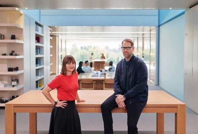 Two adults, a woman in red and a man in a blue jacket, pose behind a light wood table in a light-filled modern office space.