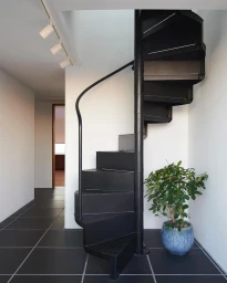 Black metal spiral staircase in a modern minimalist interior with dark floor tiles and a potted plant.