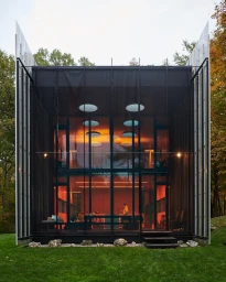 Modern glass house exterior with warm interior lighting set against a green lawn and autumn trees, photographed by Jason Schmidt.
