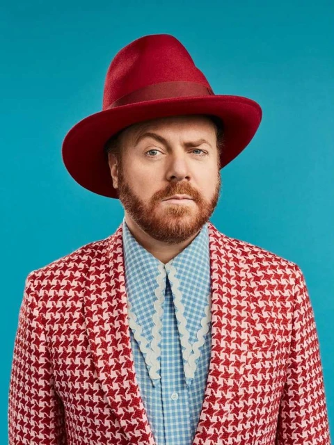Portrait of a red-bearded man in a red houndstooth blazer and a red fedora hat against a turquoise background in a photo studio.