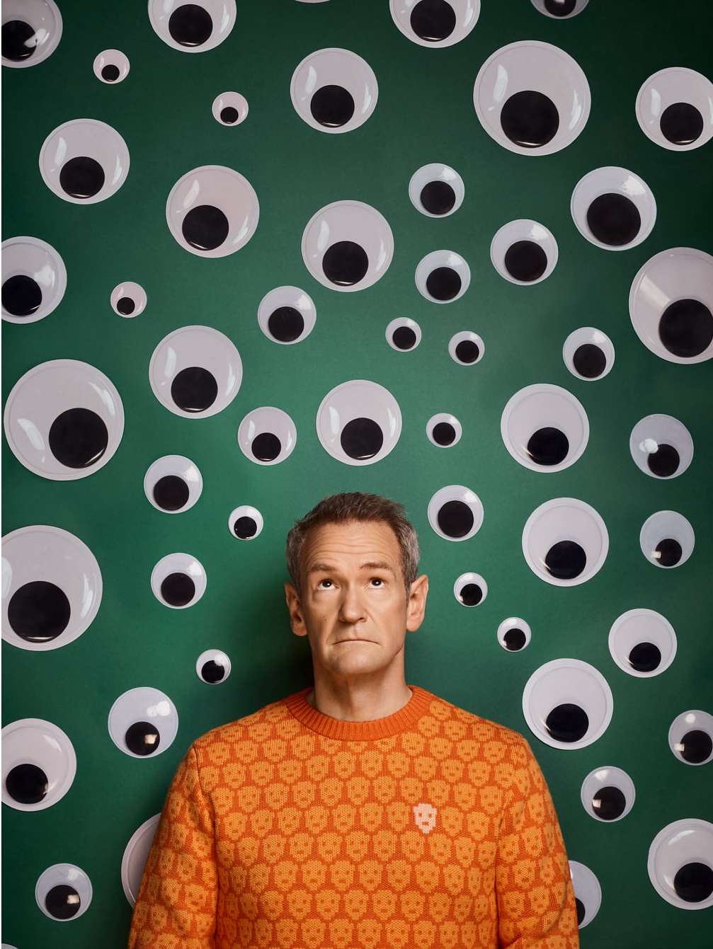An adult man in an orange skull sweater stands in front of a green wall covered in various sized googly eyes, looking directly at the camera.