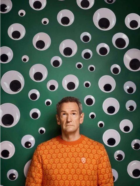 An adult man in an orange skull sweater stands in front of a green wall covered in various sized googly eyes, looking directly at the camera.