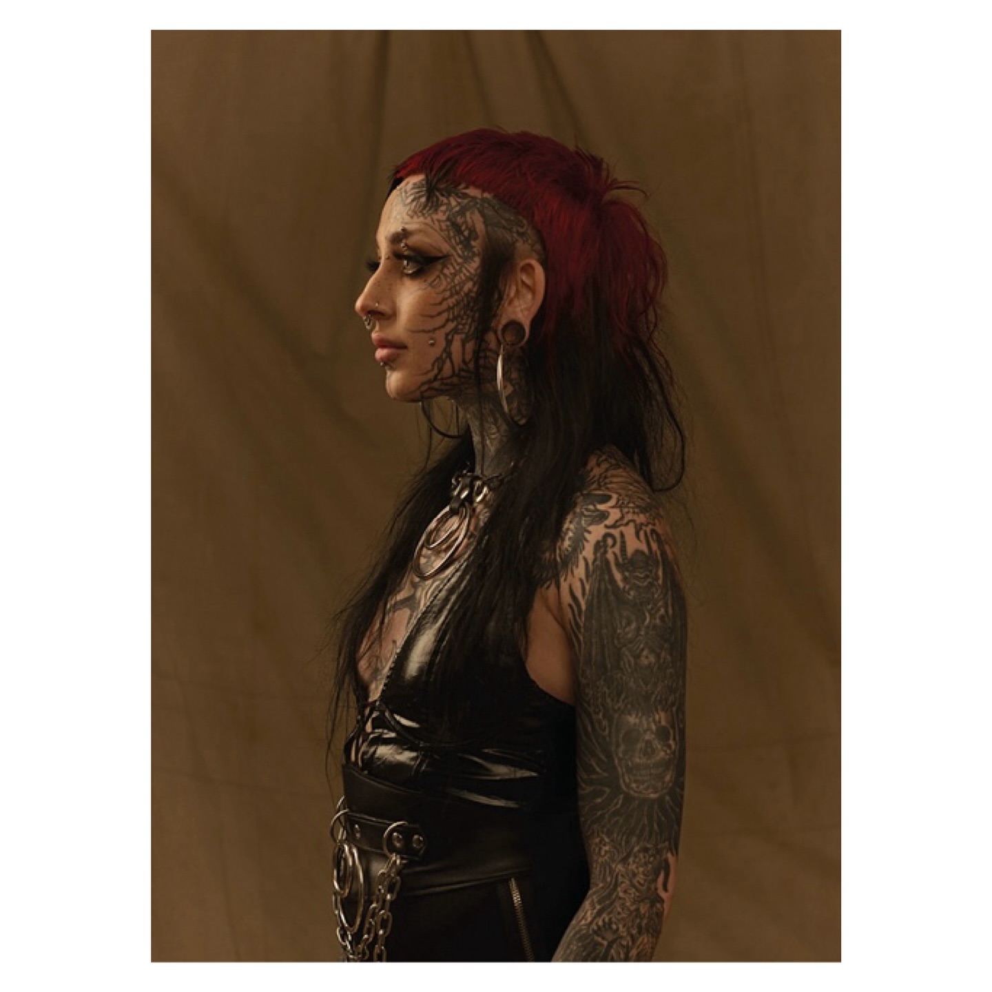 Profile portrait of a woman with tattoos, piercings, and dark red hair, wearing a black top, posing against a neutral backdrop.