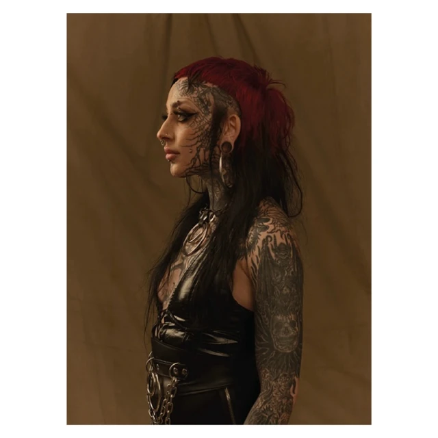 Profile portrait of a woman with tattoos, piercings, and dark red hair, wearing a black top, posing against a neutral backdrop.