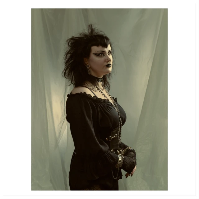 Portrait of a goth woman with dark makeup, black dress, and jewelry standing in front of a light green draped fabric background in a photo studio.