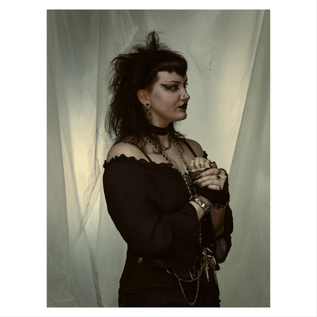 Portrait of a young adult woman with heavy black eye makeup and layered necklaces against a plain background in a studio setting.