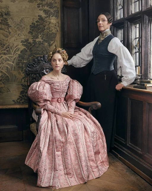 Two women pose in an indoor setting. One woman is seated in a pink gown with large sleeves, while the other stands in formal attire.
