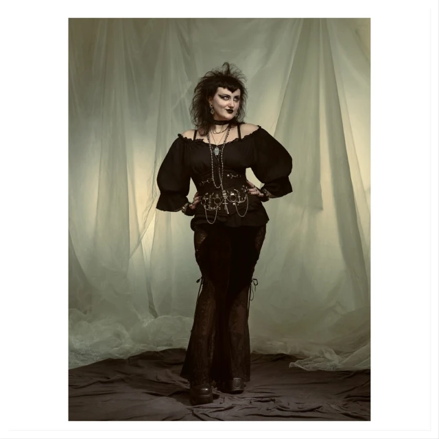 Full-length portrait of a gothic woman in black clothing with decorative chains and layered necklaces posing against a textured backdrop.
