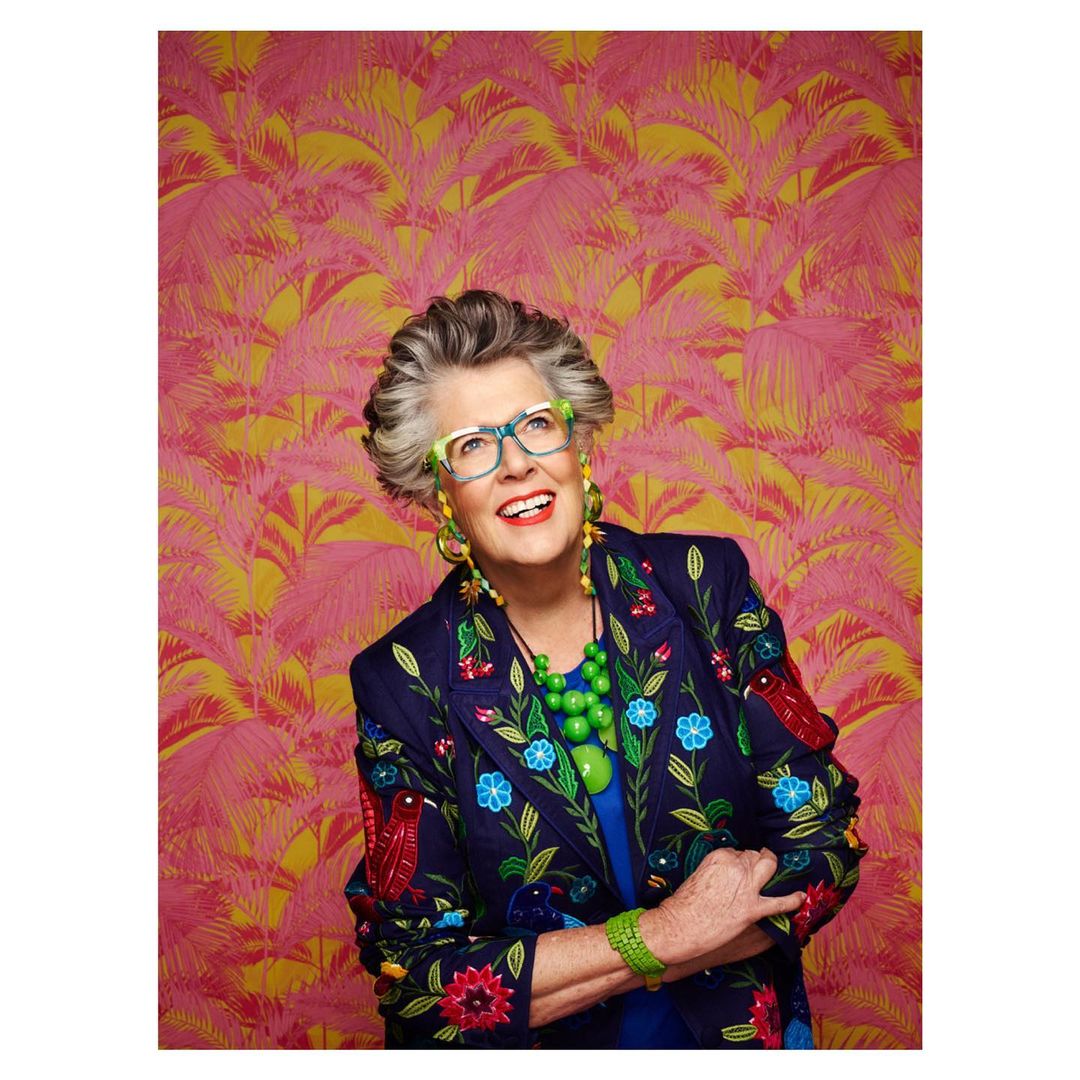 Portrait of an elderly woman with gray hair, wearing colorful glasses and an embroidered jacket, smiling against a patterned background.