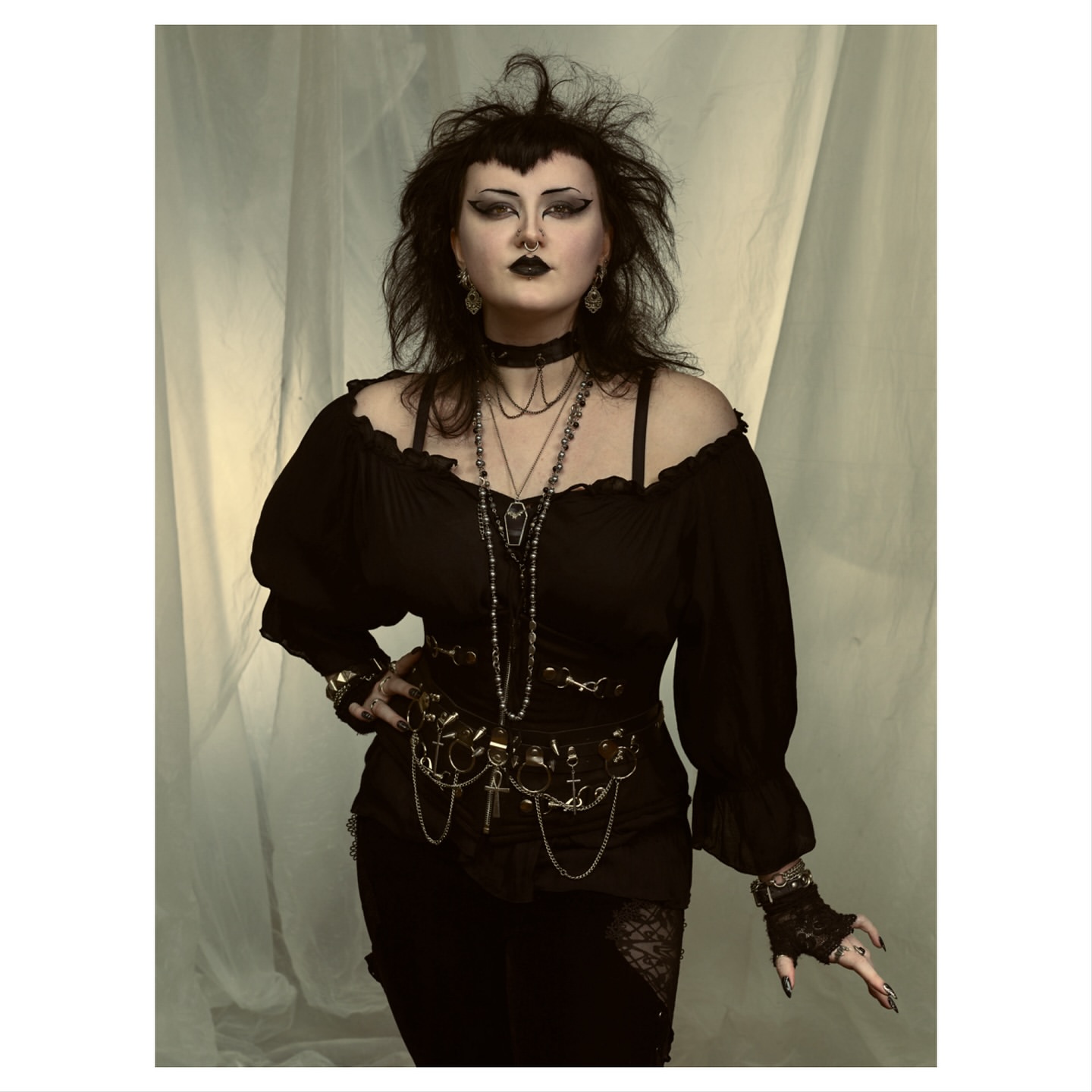 Woman with dark eye makeup and gothic clothing poses in front of a light colored backdrop, wearing jewelry and a decorative belt.