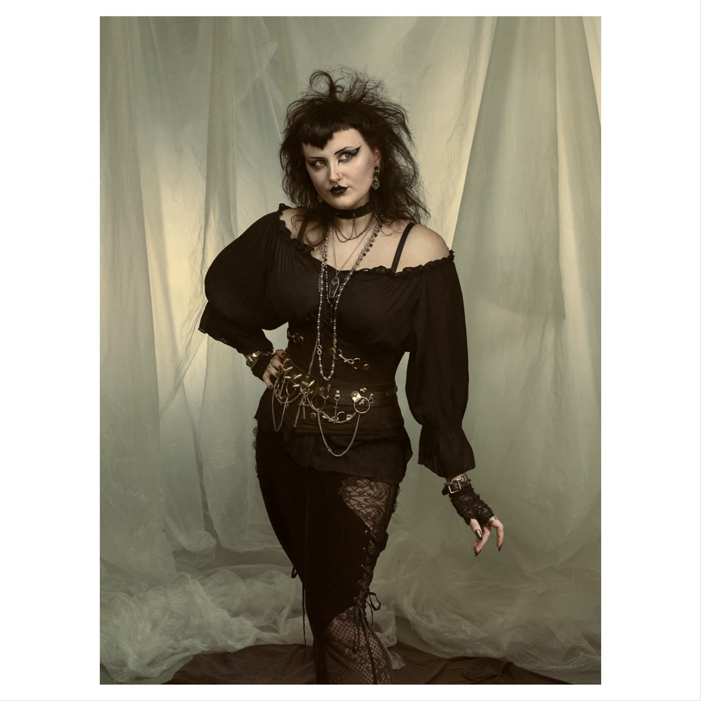 Dark-haired woman in a dark gothic-style outfit poses in front of a light-colored curtain backdrop in a studio environment.
