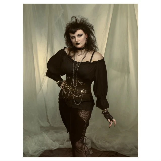 Dark-haired woman in a dark gothic-style outfit poses in front of a light-colored curtain backdrop in a studio environment.