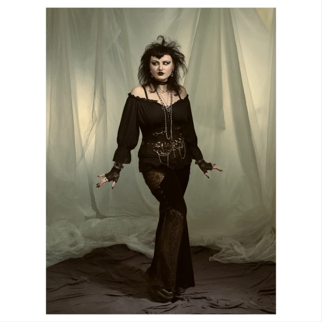 Full-length portrait of a goth woman in a black off-shoulder top and lace pants posing in front of a soft-focus backdrop with dramatic eye makeup.