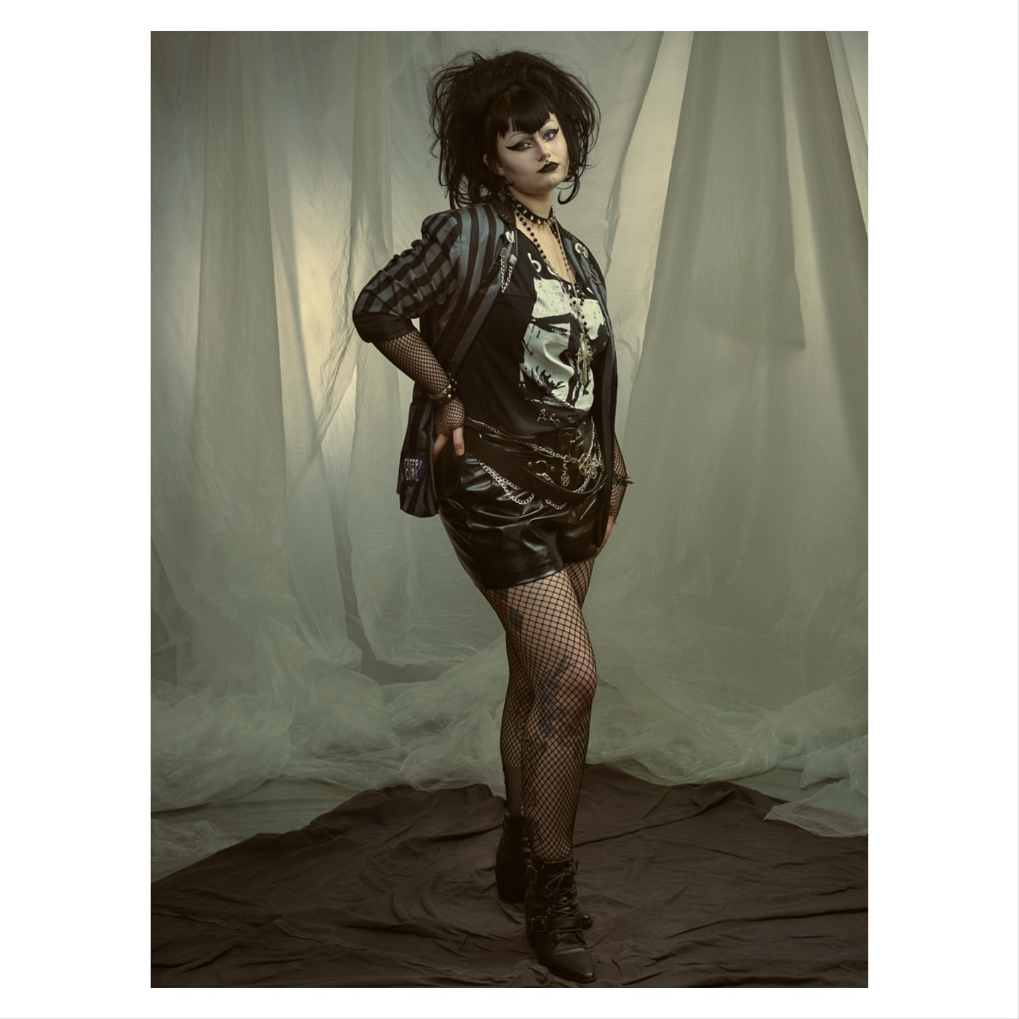 Full-length portrait of a woman with dark hair and makeup wearing shorts, fishnets, a graphic tee, blazer, and heeled boots against a studio backdrop.