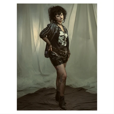 Full-length portrait of a woman with dark hair and makeup wearing shorts, fishnets, a graphic tee, blazer, and heeled boots against a studio backdrop.