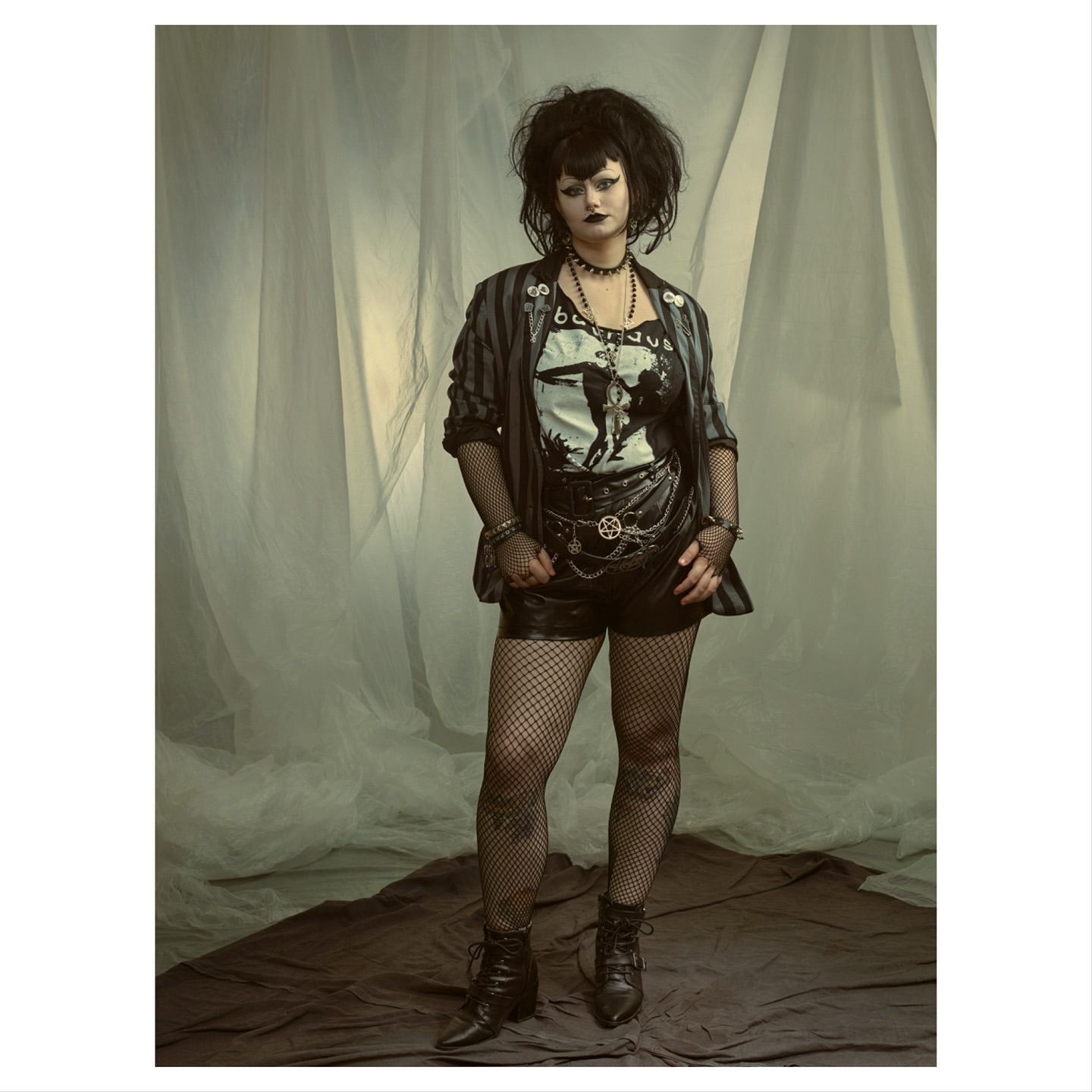 Full-length studio portrait of a woman with dark hair and alternative fashion posing in front of a sheer white and grey curtain.