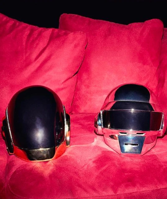 Two Daft Punk helmets, one chrome and one black, sit on a red velvet couch against a black backdrop, evoking electronic music culture.