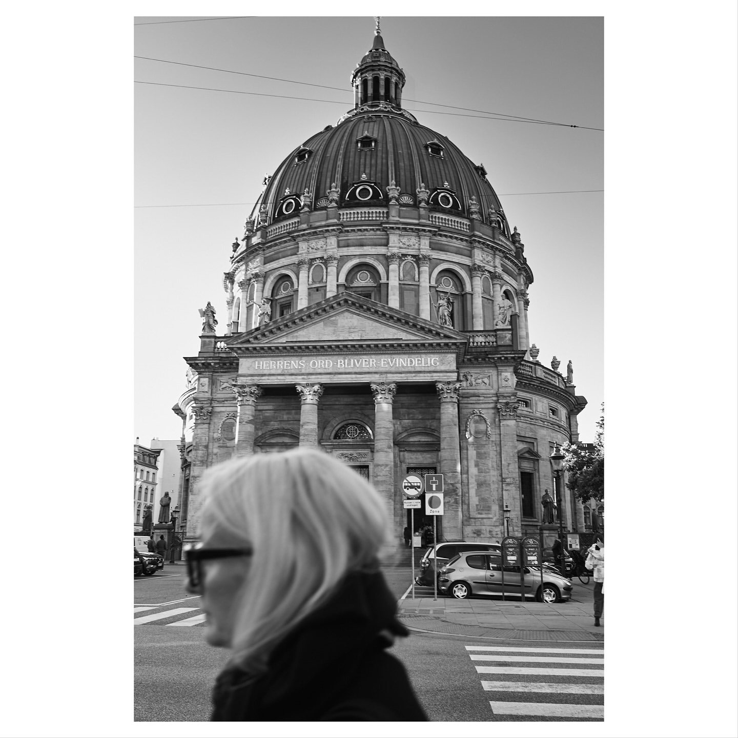 Black-and-white eye-level shot of a woman walking on a crosswalk in front of the marble dome of the Frederik's Church in Copenhagen.