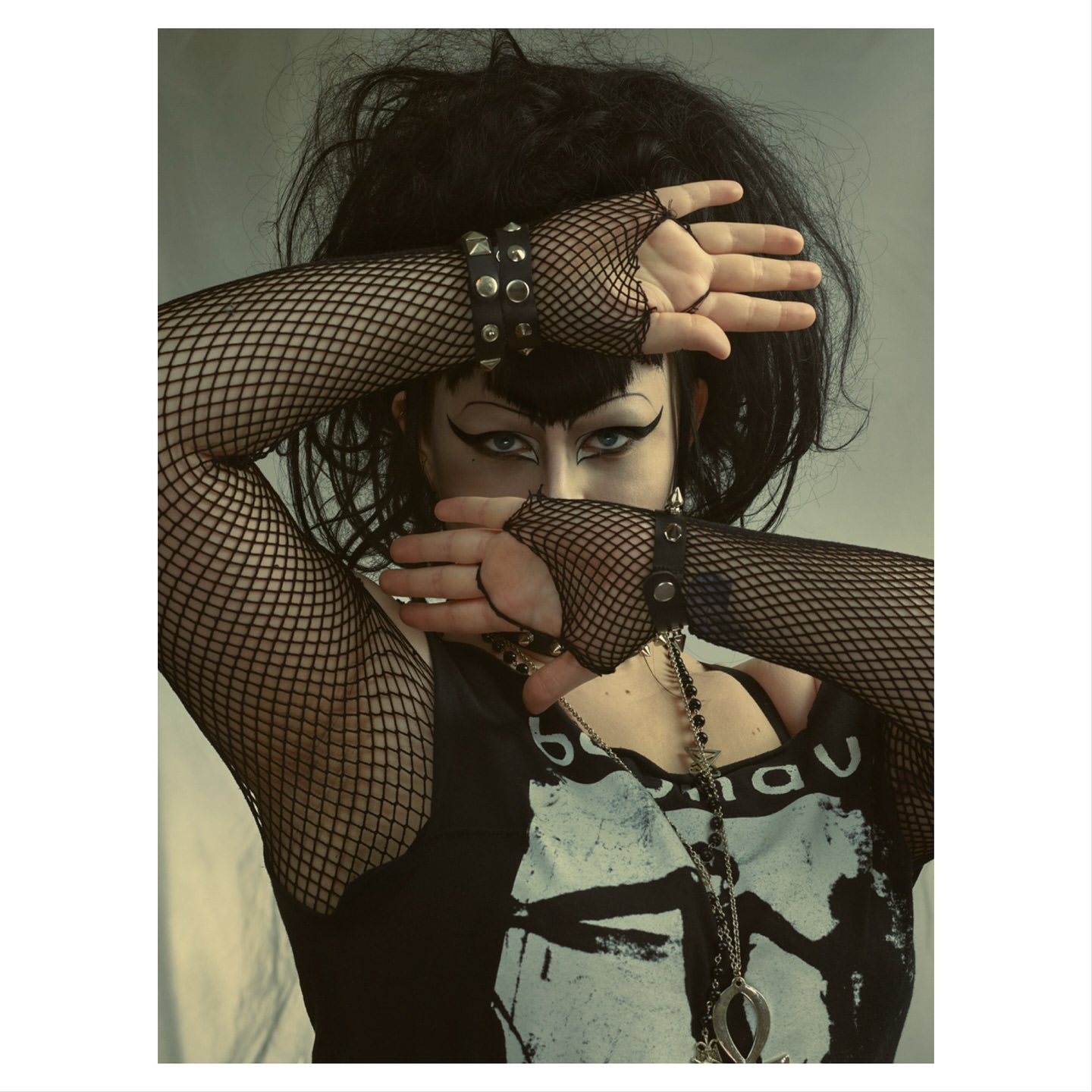 Portrait of a person with dark hair and dramatic makeup wearing fishnet gloves, their hands crossed in front of their face.