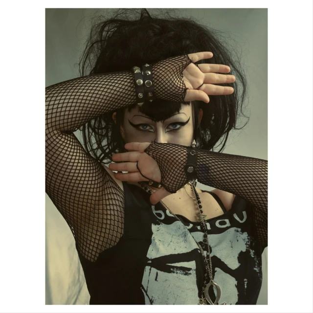 Portrait of a person with dark hair and dramatic makeup wearing fishnet gloves, their hands crossed in front of their face.