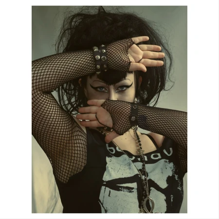 Portrait of a person with dark hair and dramatic makeup wearing fishnet gloves, their hands crossed in front of their face.