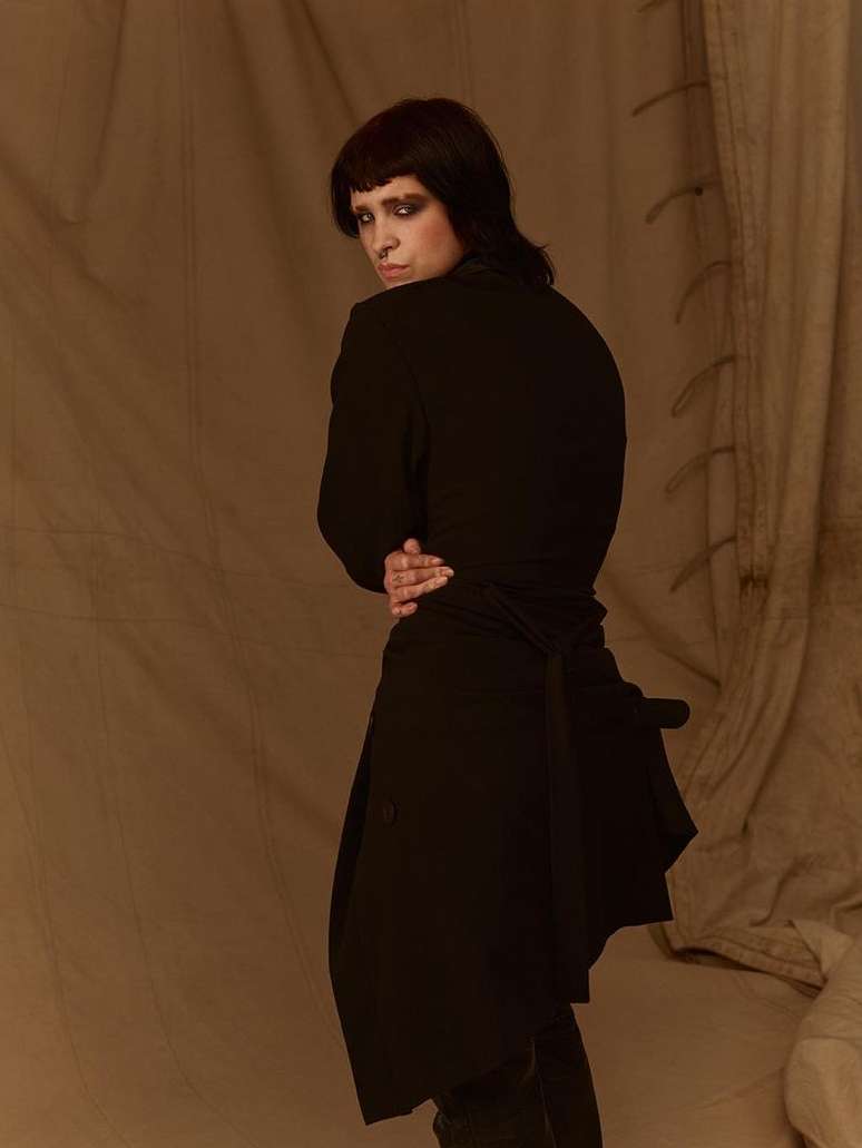 Portrait of a person with dark hair and heavy eye makeup wearing a black coat against a textured fabric backdrop