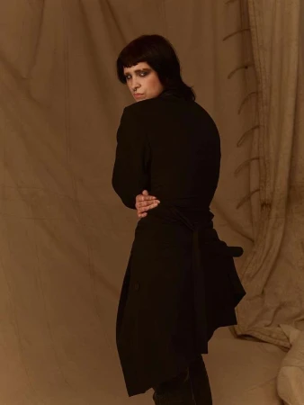 Portrait of a person with dark hair and heavy eye makeup wearing a black coat against a textured fabric backdrop