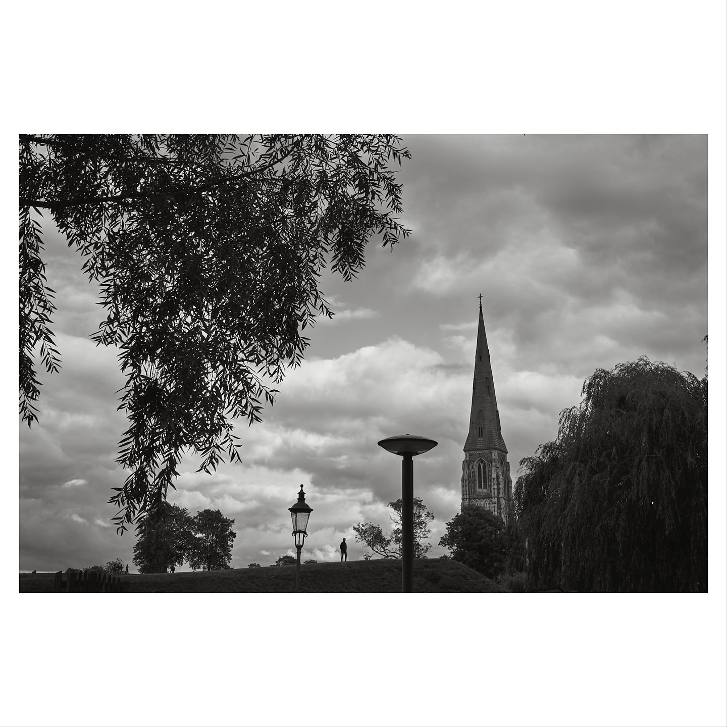 Black and white landscape featuring a distant church spire, street lamps, trees, and a cloudy sky, evoking a contemplative mood.