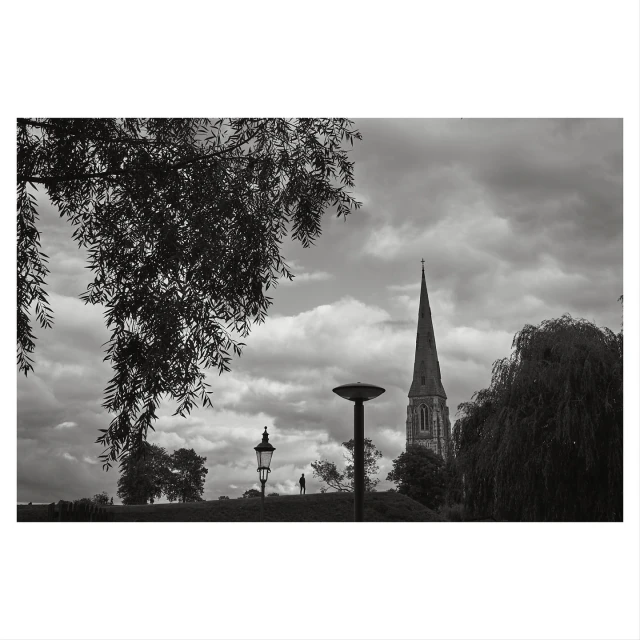 Black and white landscape featuring a distant church spire, street lamps, trees, and a cloudy sky, evoking a contemplative mood.
