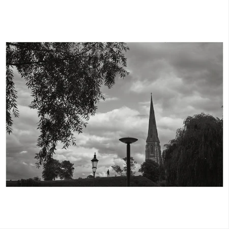 Black and white landscape featuring a distant church spire, street lamps, trees, and a cloudy sky, evoking a contemplative mood.