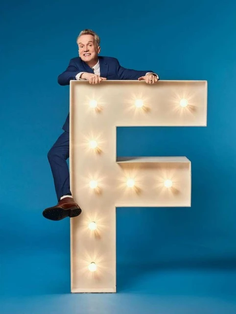 Adult male in a blue suit posing behind a large illuminated letter F against a plain blue background, creating a playful studio portrait.