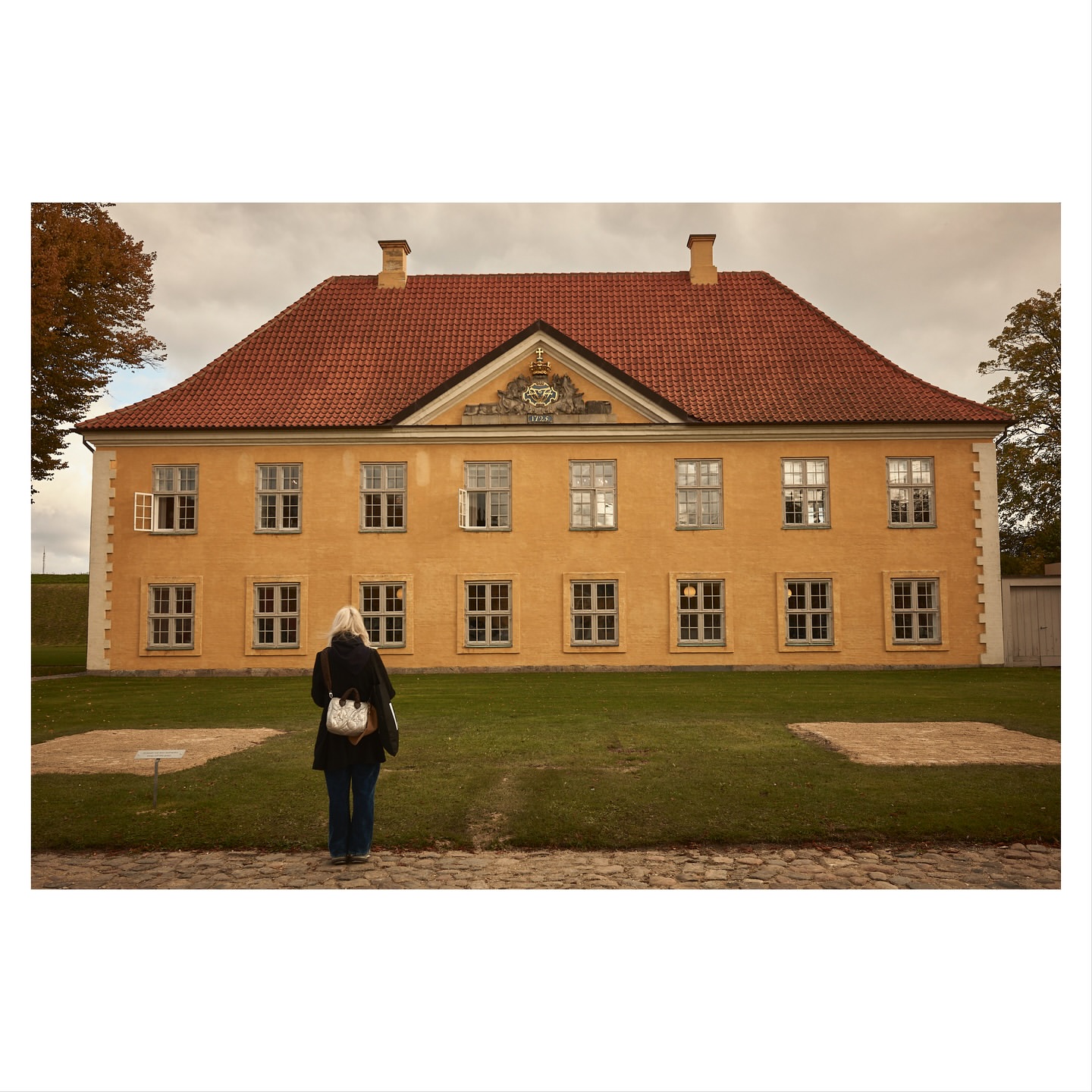 Woman stands on brick path, facing a yellow building with a red roof on a sunny day with green grass in front of building.