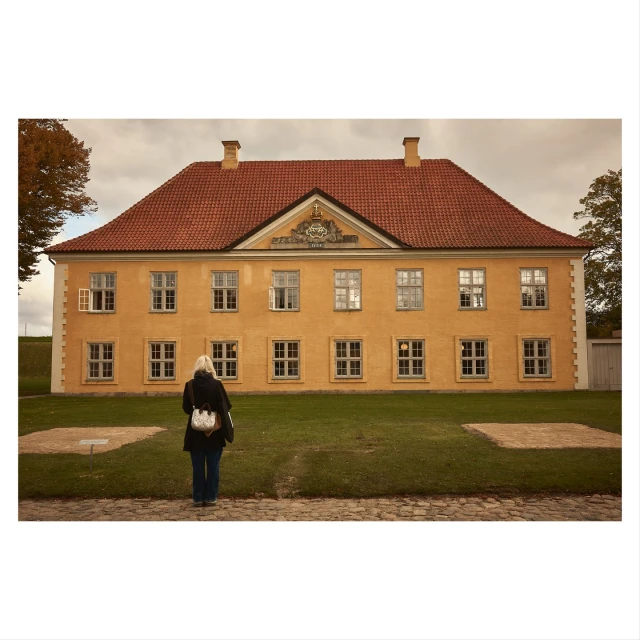Woman stands on brick path, facing a yellow building with a red roof on a sunny day with green grass in front of building.