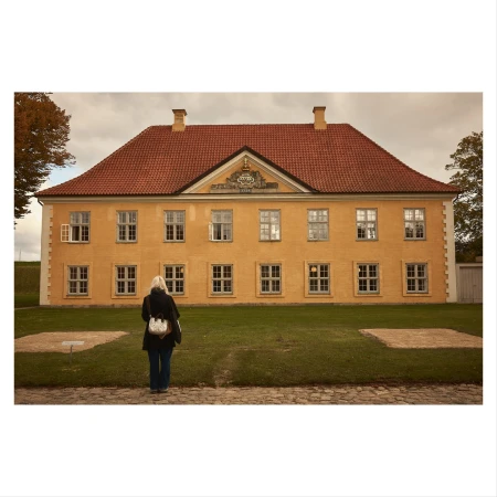 Woman stands on brick path, facing a yellow building with a red roof on a sunny day with green grass in front of building.