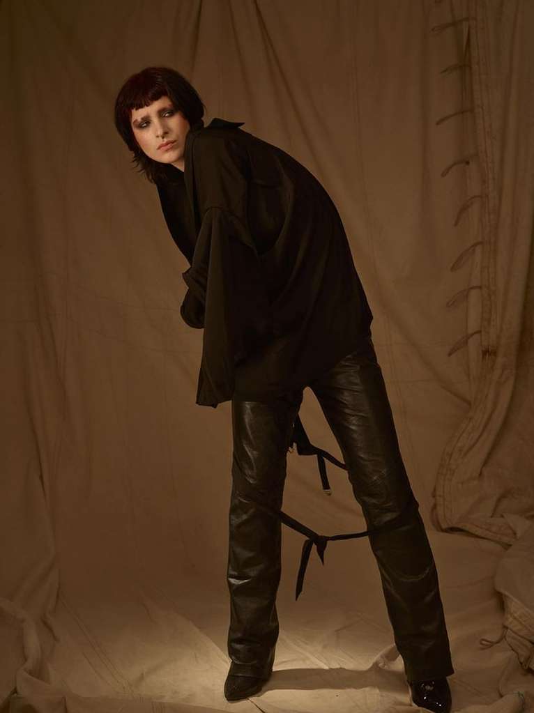 A person in a black top and black leather pants with straps stands in a studio with a neutral backdrop, fashion portrait, medium shot.
