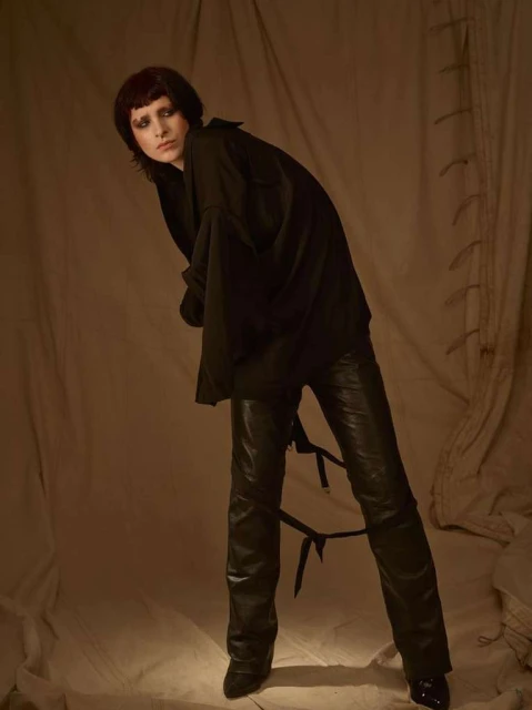 A person in a black top and black leather pants with straps stands in a studio with a neutral backdrop, fashion portrait, medium shot.