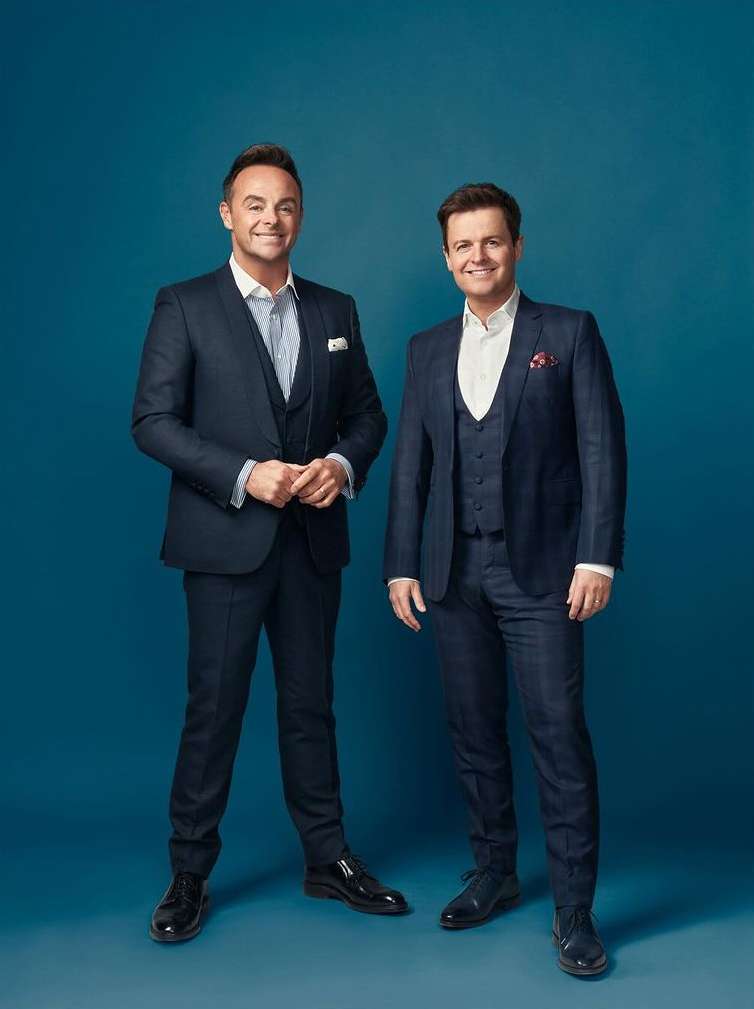 Two adult males in matching blue suits pose indoors against a solid blue backdrop, smiling and looking directly at the camera.