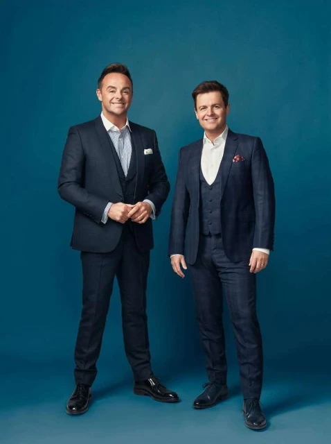 Two adult males in matching blue suits pose indoors against a solid blue backdrop, smiling and looking directly at the camera.
