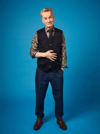 Elderly man in a patterned shirt, black vest, and trousers standing against a vibrant blue studio backdrop