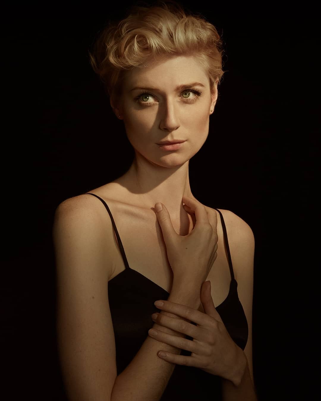 Elegant portrait of a blonde woman with fair skin against a black backdrop. She wears a black tank top and has her hands gently posed.