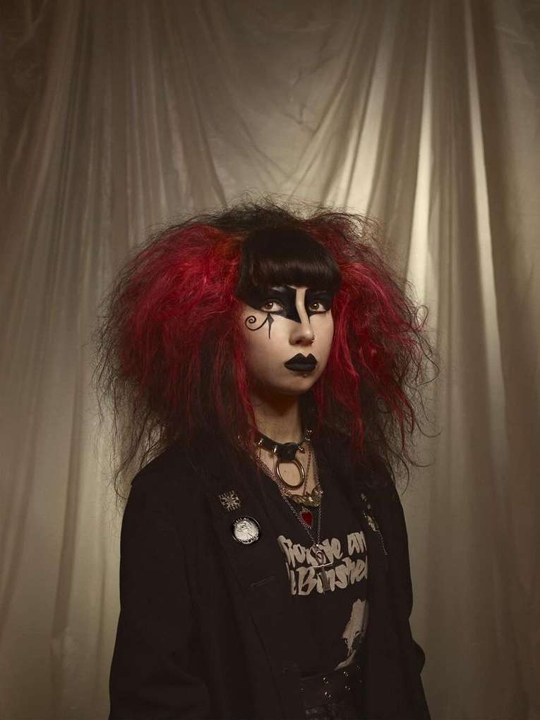 Portrait of a woman with goth makeup and vibrant red and black hair, wearing a black jacket, against a textured beige backdrop.